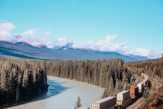 A cargo train travels alongside a lush river through a mountainous forest landscape on a clear day.