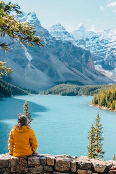 A woman in a yellow jacket sits by a scenic lake, admiring snow-covered mountains.