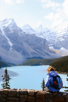 A woman in a blue jacket sits on a stone wall overlooking the serene Moraine Lake and mountains.