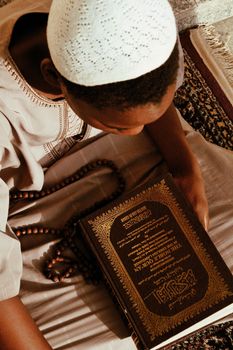 From above of crop black male wearing robe sitting on floor with Quran and rosary beads while praying