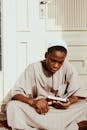Concentrated African male in Islamic prayer cap and robe reading Koran sitting on floor against door