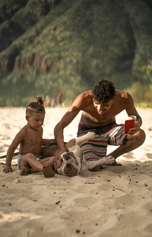 Full body of happy ethnic male in swimwear sitting on sandy beach with son and playing with dog