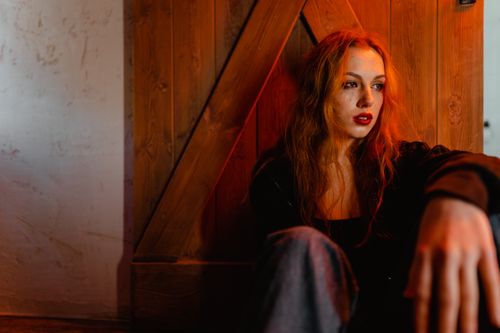 Free Red-haired woman sitting by a wooden door in warm, moody lighting expressing emotion. Stock Photo