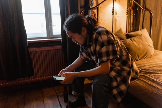A young man with a contemplative expression sits on a bed looking at his phone inside a cozy room.
