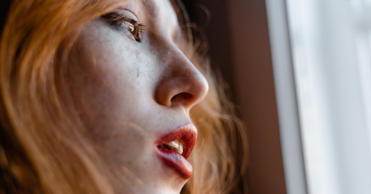 A young woman with red hair looks thoughtfully out a window, capturing an emotional moment.