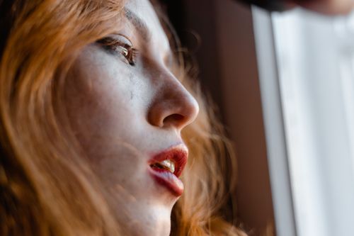 Free A young woman with red hair looks thoughtfully out a window, capturing an emotional moment. Stock Photo