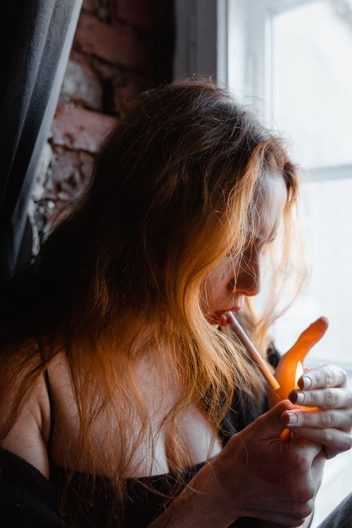 Free A woman lights a cigarette by a window, creating a dramatic and moody scene. Stock Photo