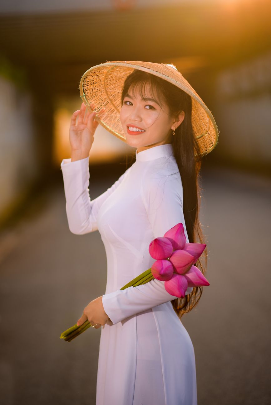Woman in ao dai and conical hat smiles while holding lotus flowers at sunset.
