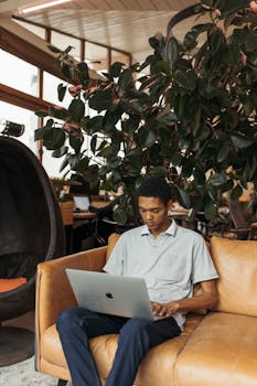 A young man working on a laptop in a modern indoor workspace with plants.