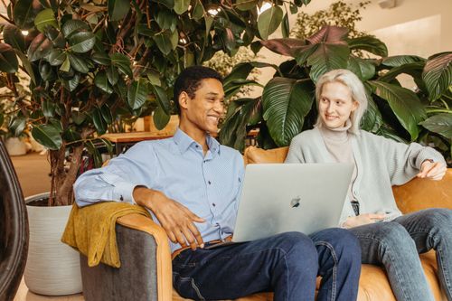 Free Two colleagues sharing ideas and smiles while working on a laptop in a lively office setting. Stock Photo
