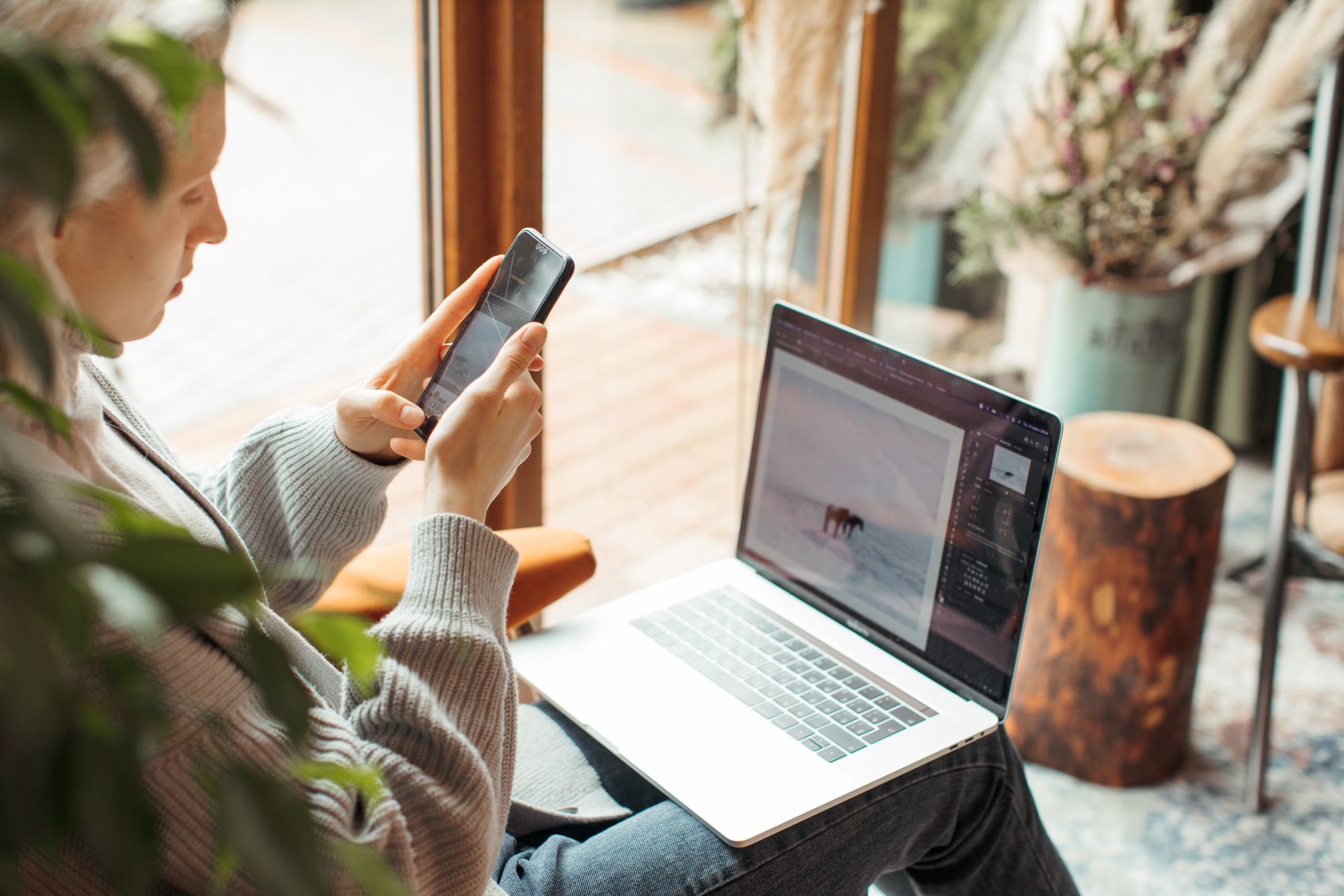 A woman using her smartphone and laptop for remote work indoors, capturing a cozy workspace.