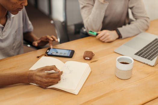 Two colleagues brainstorming with a notebook, laptop, and coffee on a wooden table.