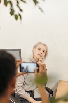 Candid shot of a woman posing thoughtfully while being photographed with a smartphone in an office setting.
