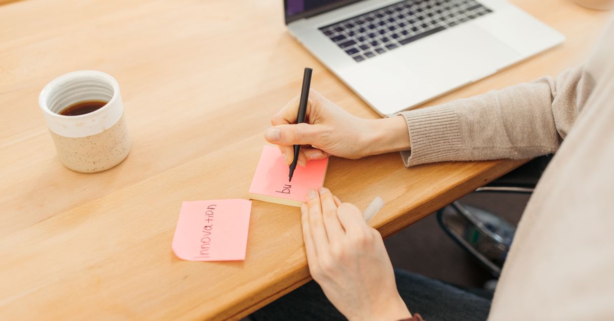 person writing on a sticky note next to a laptop with a coffee cup, overhead angle