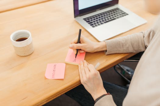 Person writing on a sticky note next to a laptop, showcasing a work or study setup with coffee.