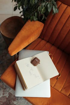 A stylish workspace featuring a notebook and laptop on a plush orange armchair, perfect for cozy work sessions.