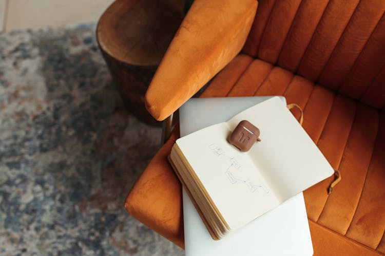 An Open Notebook With  Airpods Case On An Orange Sofa