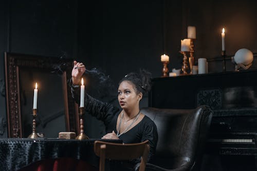 Woman in Black Long Sleeve Shirt Sitting and Holding a Match Stick In Front of Lighted Candles