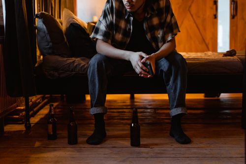 Free A man sits on a bed holding a cellphone, surrounded by beer bottles, creating a moody atmosphere. Stock Photo