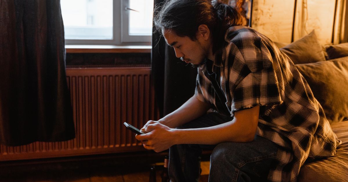 Um homem com uma camisa xadrez usando um smartphone enquanto está sentado em um quarto acolhedor.