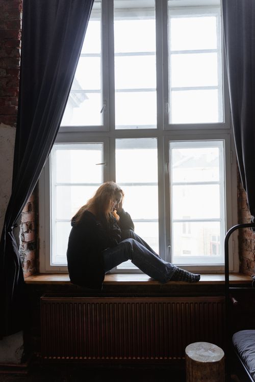 Free A woman sits thoughtfully on a window sill, gazing outside with curtains open. Stock Photo