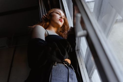 Free A woman gazes out of a window, deep in thought in a softly lit room. Stock Photo