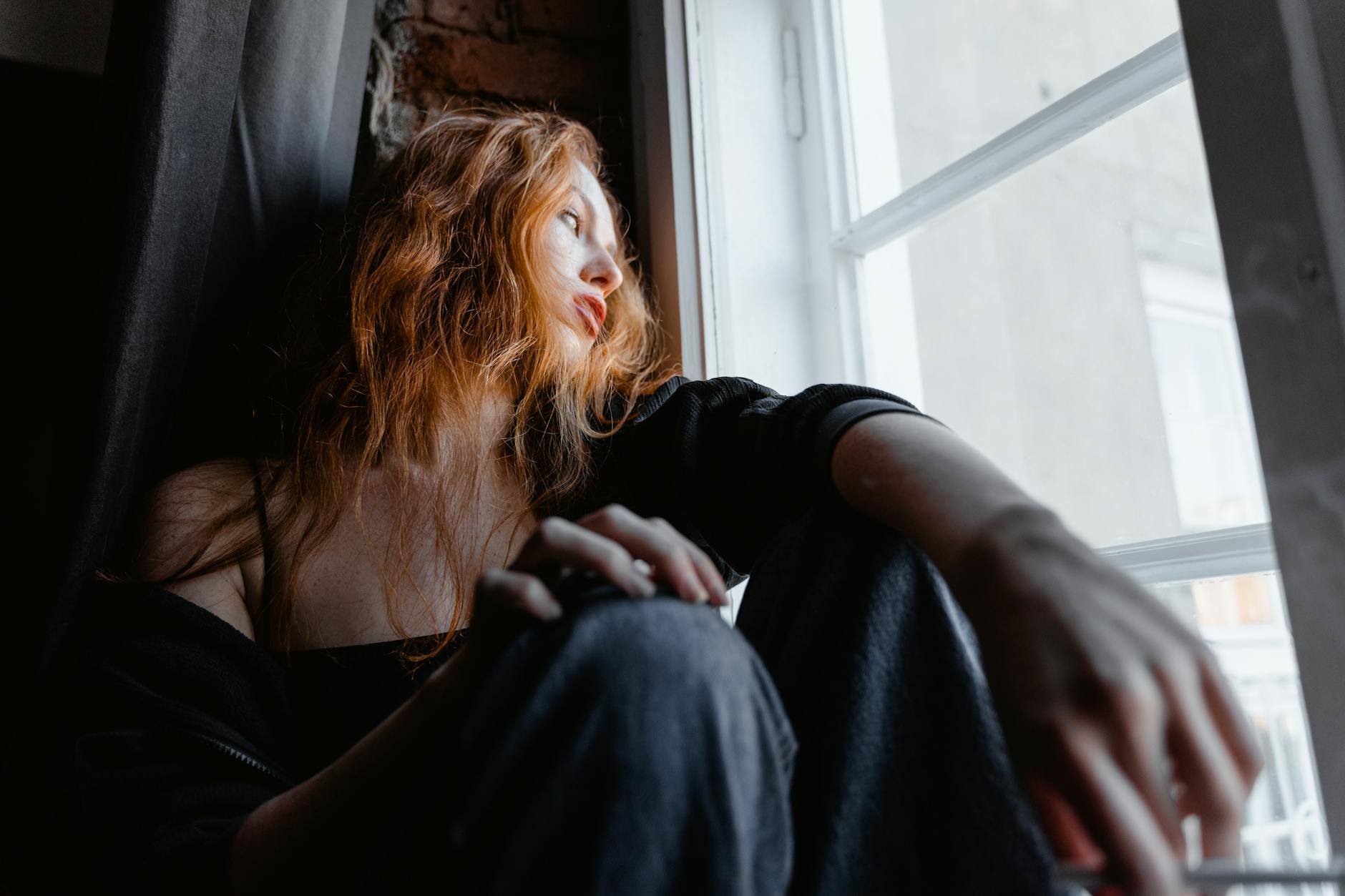 A woman with red hair sits by a window, deep in thought, conveying emotion and reflection.