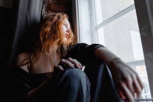 Free A woman with red hair sits by a window, deep in thought, conveying emotion and reflection. Stock Photo