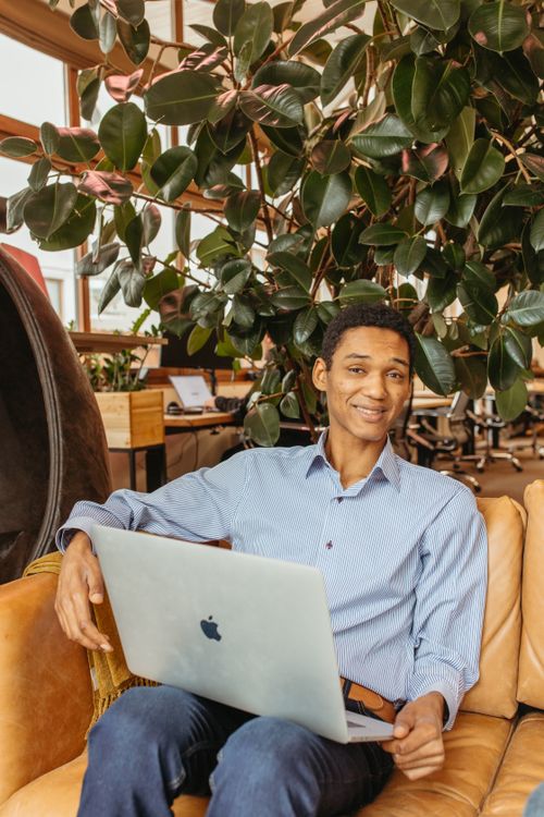 Free Adult man sitting on a couch with a laptop, surrounded by indoor plants in an office setting. Stock Photo
