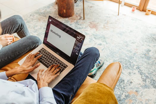 Young adult typing on a laptop for marketing work, seated indoors in a casual setting.