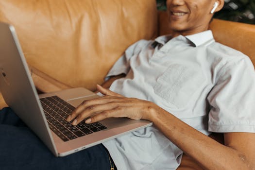 Man with AirPods works on laptop while relaxing on a couch, embodying modern remote work lifestyle.