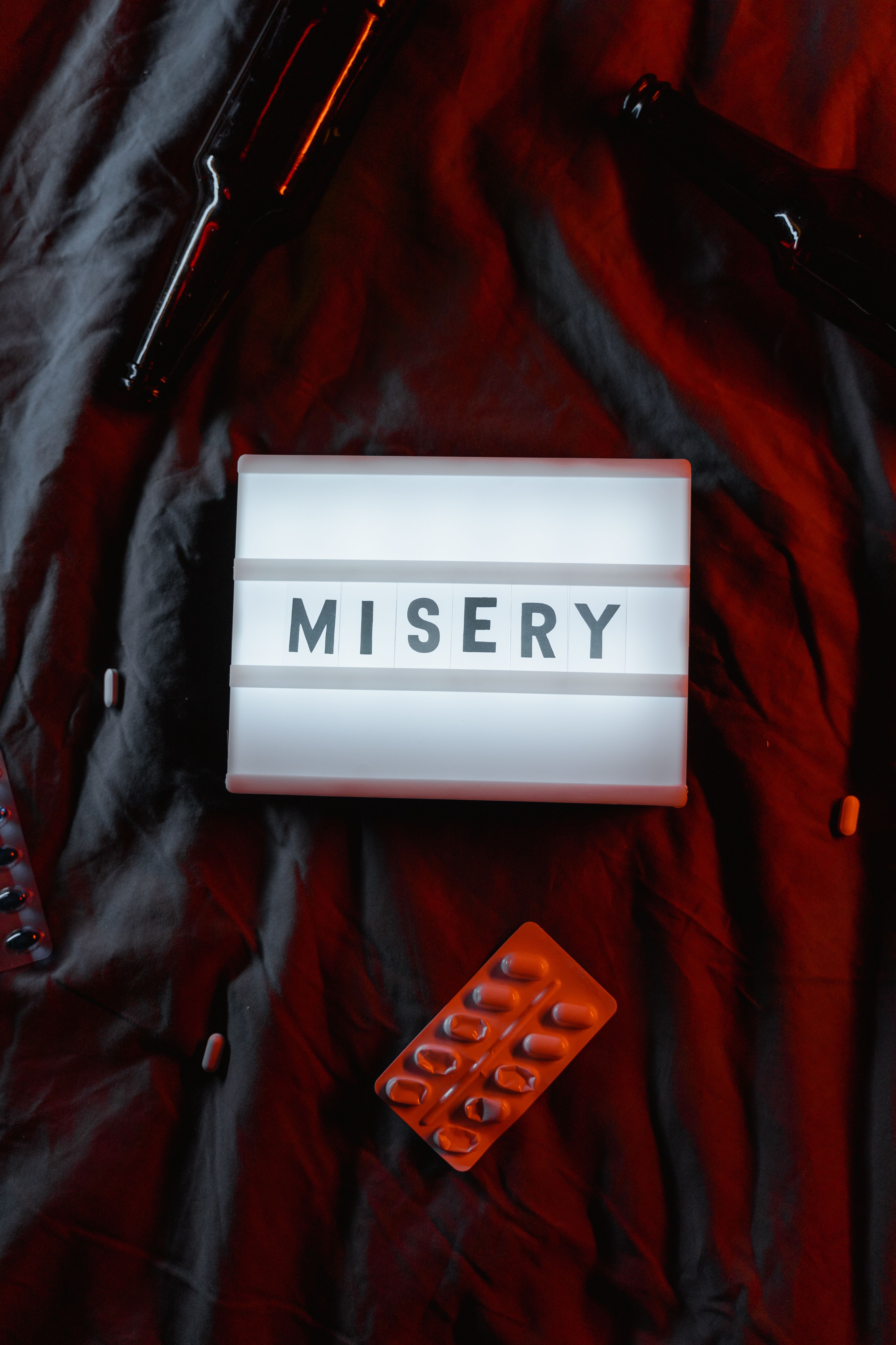 Flat lay composition with word 'Misery', pills, and empty beer bottles on red fabric.