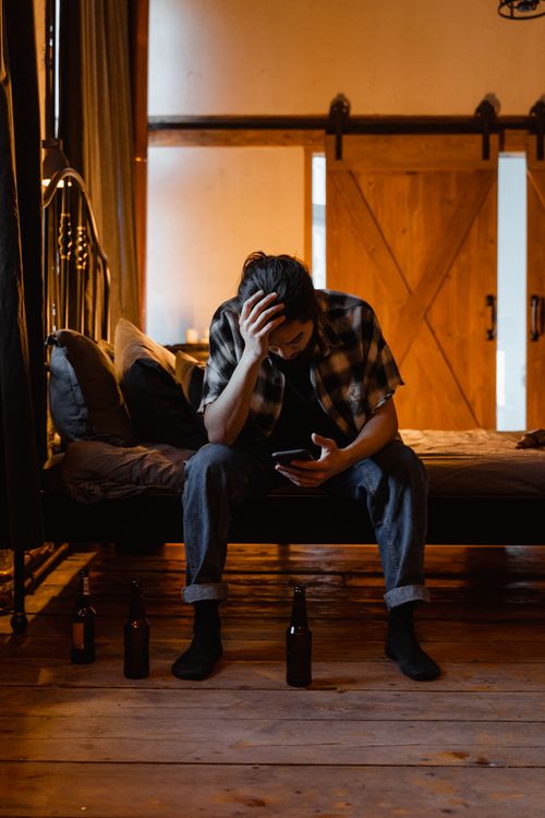 Free Young man in a flannel shirt sitting on bed, looking at phone, surrounded by bottles, in a dimly lit room. Stock Photo