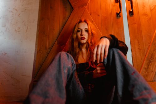 Free A woman with long hair sits pensively indoors against a wooden door in warm lighting. Stock Photo