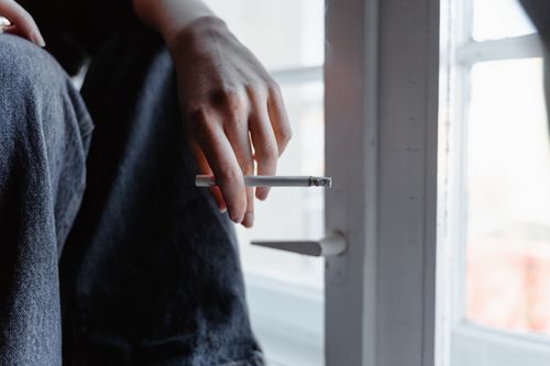 Free A person's hand holding a cigarette by a window, suggesting thoughtful solitude. Stock Photo