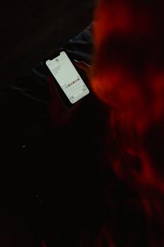A person texting on a smartphone in a dark room with red lighting.