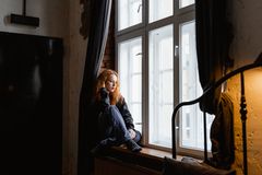 A thoughtful woman sits by a window in a cozy room, deep in contemplation.