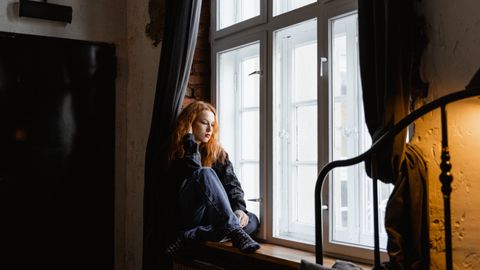 A thoughtful woman sits by a window in a cozy room, deep in contemplation.