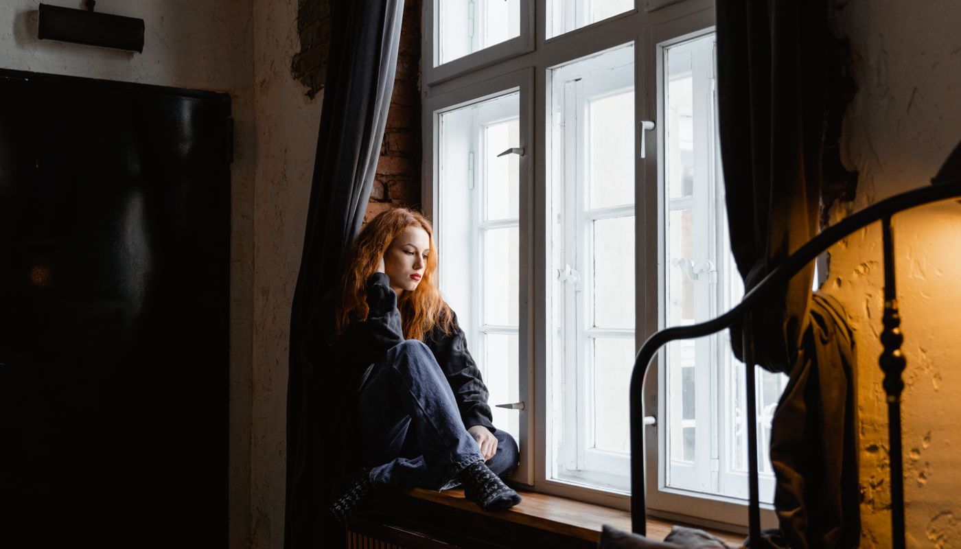 A woman sits on a windowsill with her knees drawn up, looking down in thought, in a room with bare brick walls and a heavy curtain