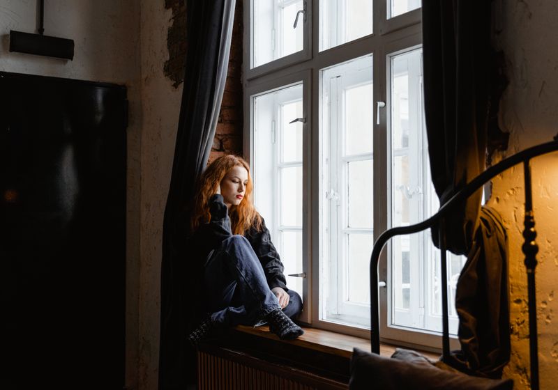 A woman sits on a windowsill with her knees drawn up, looking down in thought, in a room with bare brick walls and a heavy curtain
