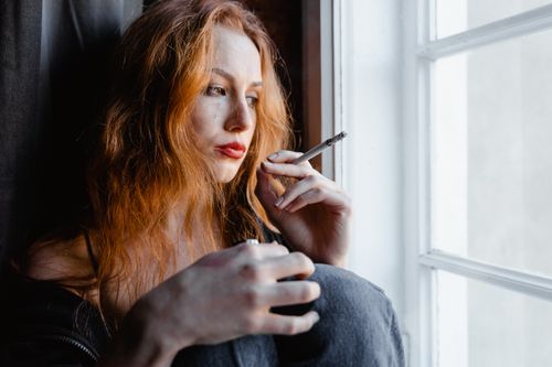 Free Pensive young woman with red hair holding a cigarette by a window, reflecting indoors. Stock Photo