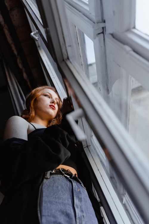 Free A young woman stands by a window, reflecting on her thoughts indoors. Stock Photo