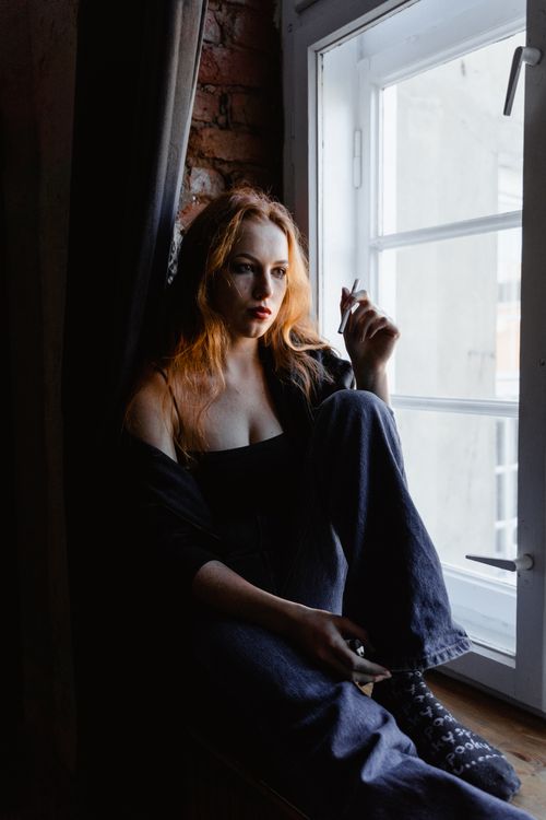 Free Pensive woman sits by a window, holding a cigarette, indicating deep thoughts or emotions. Stock Photo