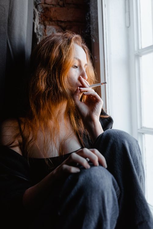Free Pensive woman with long red hair smokes by a window, enveloped in moody lighting. Stock Photo