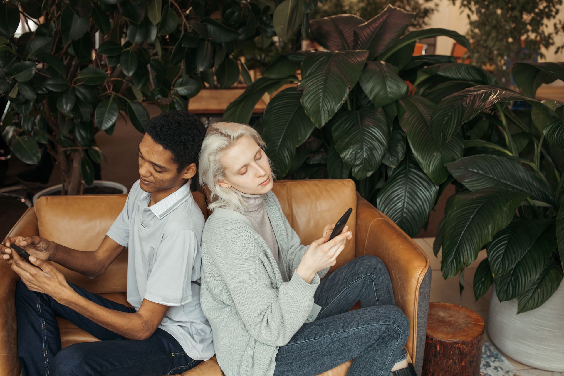 A multicultural couple sits back to back, focused on their mobile phones in a cozy indoor setting.
