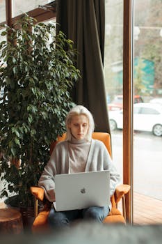 A woman focused on her laptop while seated in a cozy cafe, next to a large window with a street view.