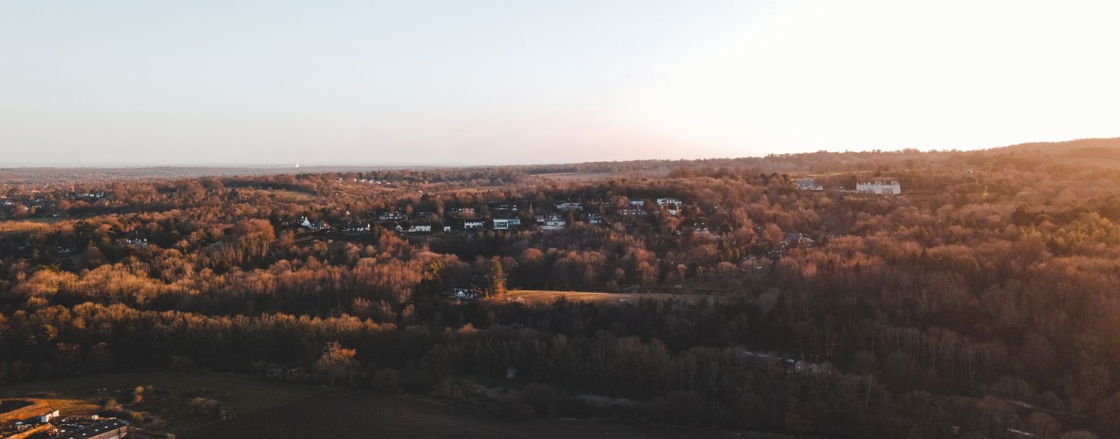 A scenic aerial shot over the English countryside at sunset showcasing houses and a vast forest.