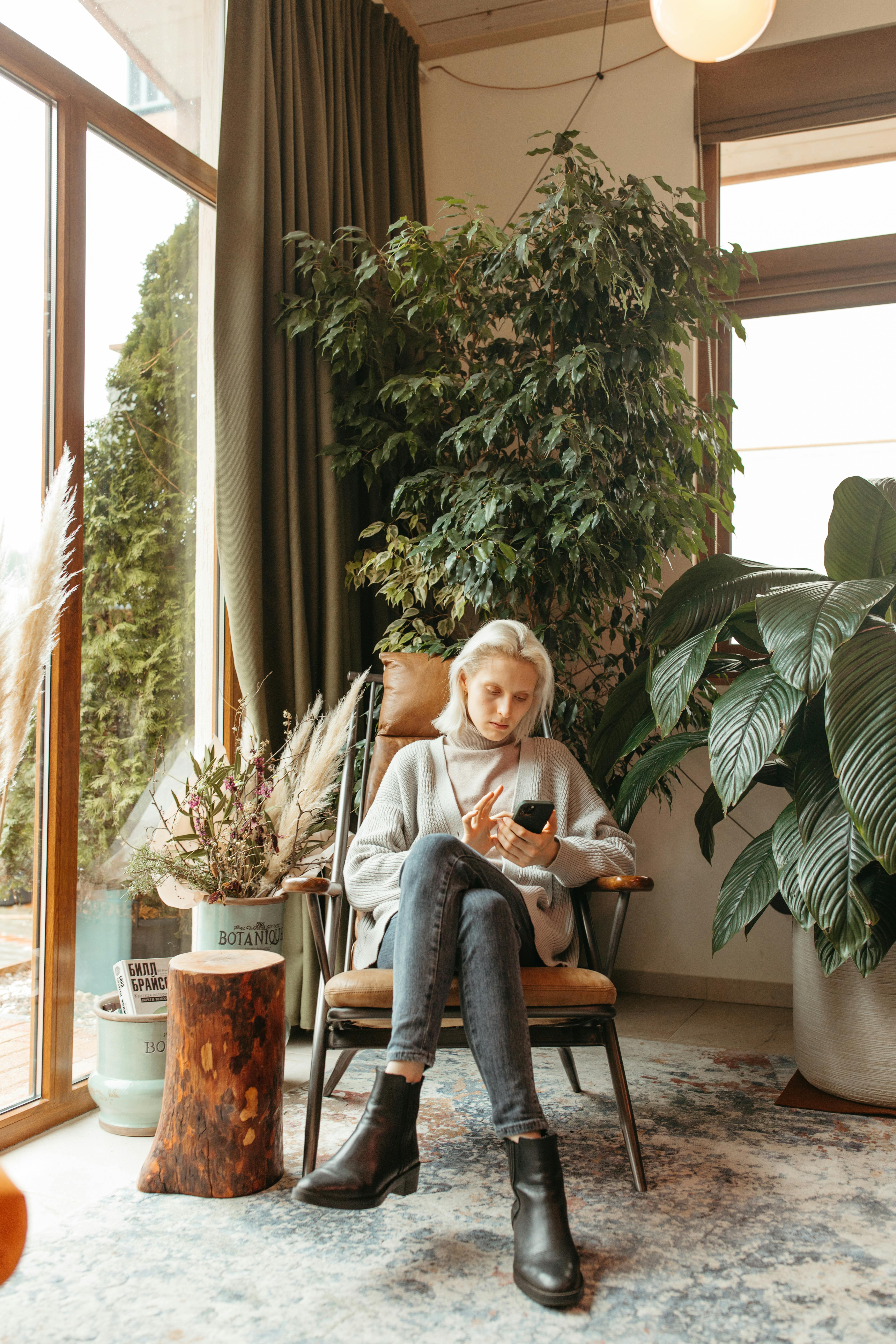 Blonde woman sitting in cozy room checking smartphone surrounded by indoor plants.