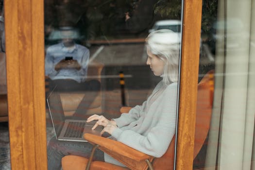 A woman with white hair works on her laptop inside a cozy cafe, seen through a window.