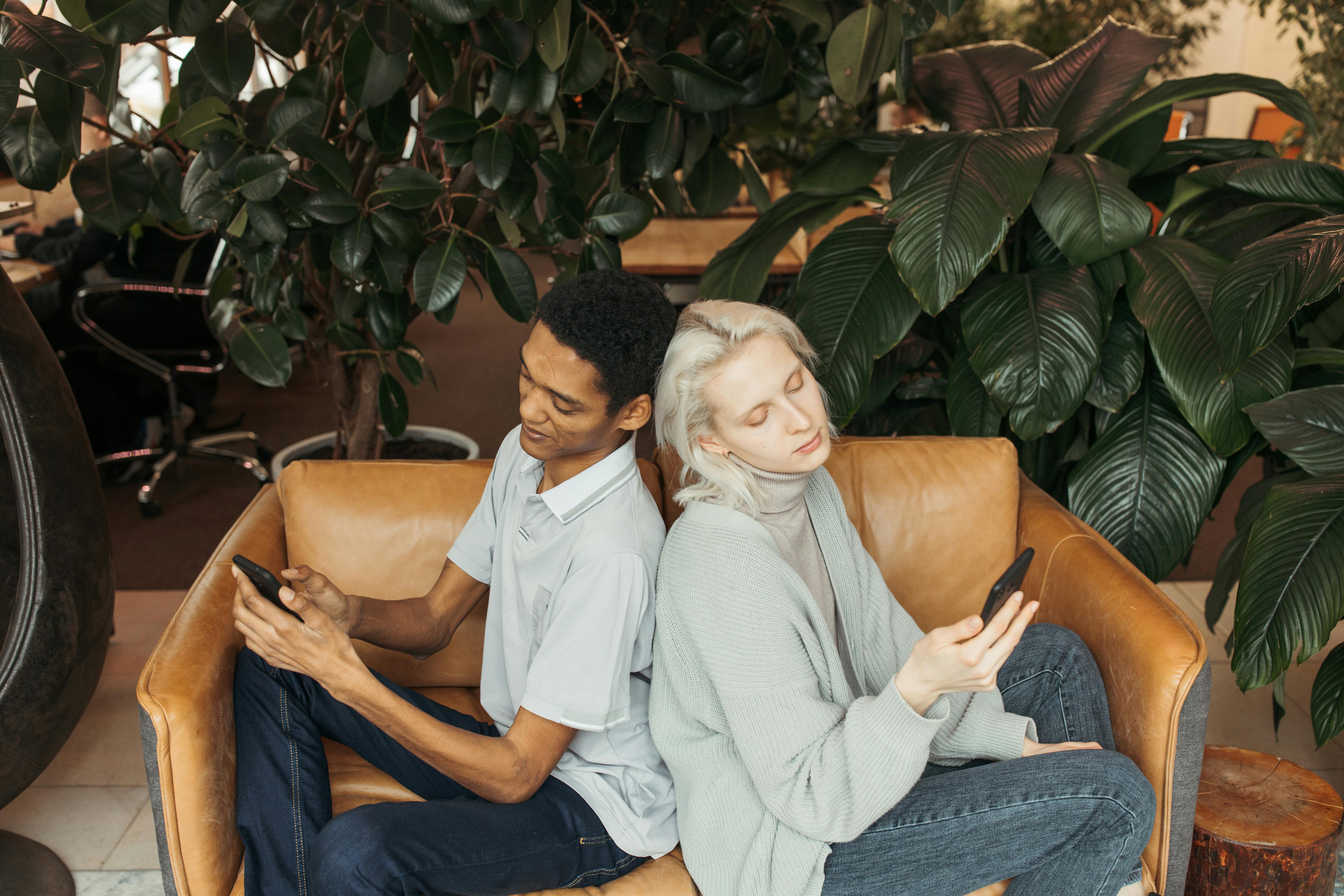 Two adults sitting back to back on a sofa, engaged with their phones, surrounded by indoor plants.